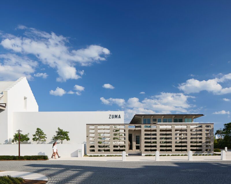 A person walks past the modern, minimalist facade of a building labeled "ZUMA" in Alys Beach under a clear blue sky dotted with clouds, capturing the serene vibe and architectural Art of the Drive along 30A.