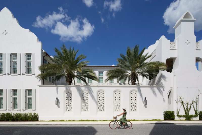 A person rides a bicycle past a white building with palm trees and arched windows in Alys Beach, showcasing the coastal charm often seen along Cars of 30A, under a blue sky with scattered clouds.
