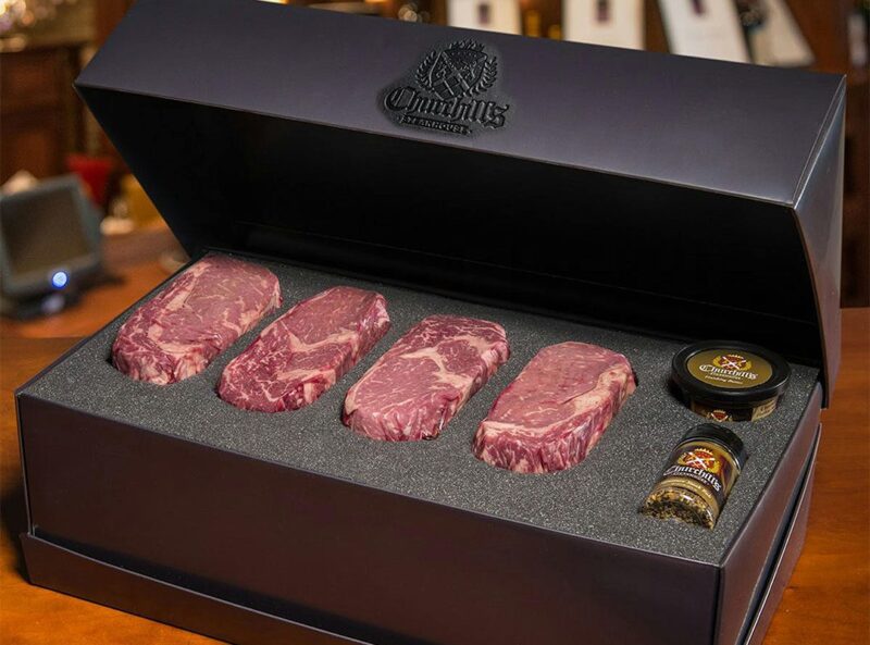 Featured in the duPont REGISTRY Father's Day Gift Guide, four raw marbled steaks and two seasoning containers are arranged in a sleek black gift box with foam padding.