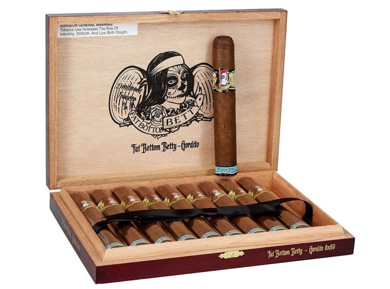 An open wooden box displaying twelve cigars with labels, featuring artwork of a woman and “Fat Bottom Betty” text on the inside lid. One cigar is propped on the edge—perfect for the duPont REGISTRY Father’s Day Gift Guide.