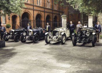 Six men in suits stand beside four vintage cars parked in a courtyard with red brick buildings and stone pavement in the background.