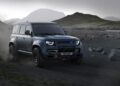 A dark blue Land Rover Defender Refreshed: Here's What’s New For 2026, driving off-road on a rocky, dusty terrain with mountains in the background under a cloudy sky.