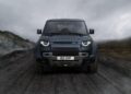 A dark blue Land Rover Defender Refreshed: Here's What’s New For 2026 is parked on a rugged dirt road, with mountains and cloudy skies in the background.
