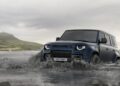 A blue Land Rover Defender Refreshed: Here's What’s New For 2026, drives through a shallow river with water splashing around its wheels; mountains and a cloudy sky are visible in the background.
