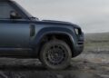 Front side view of a blue Land Rover Defender Refreshed parked on rocky terrain, with mountains in the background under a cloudy sky—here’s what’s new for 2026.