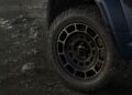 Close-up of a rugged off-road tire and wheel on a dark-colored Land Rover Defender Refreshed: Here's What’s New For 2026, parked on uneven, rocky terrain.