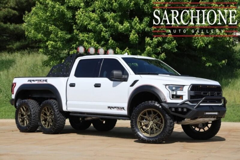 A white six-wheel Ford Raptor pickup truck with off-road modifications is parked on pavement in front of green trees. This unique garage creation is featured as part of the "Sarchione Auto Gallery," whose logo appears in the top right corner.
