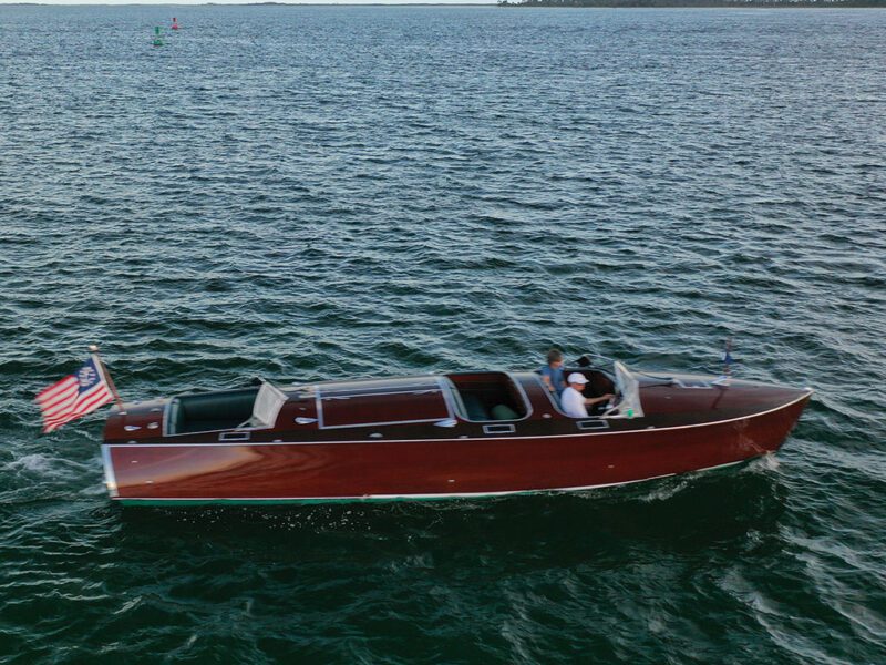 A mahogany wooden boat with an American flag glides across a large body of water, carrying two people seated inside.