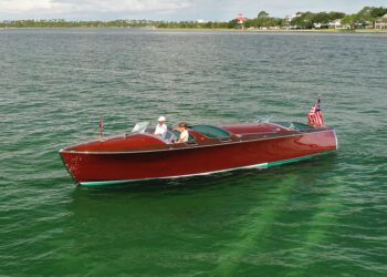 A classic mahogany wooden boat with two people onboard cruises across calm green water, displaying an American flag at the stern.