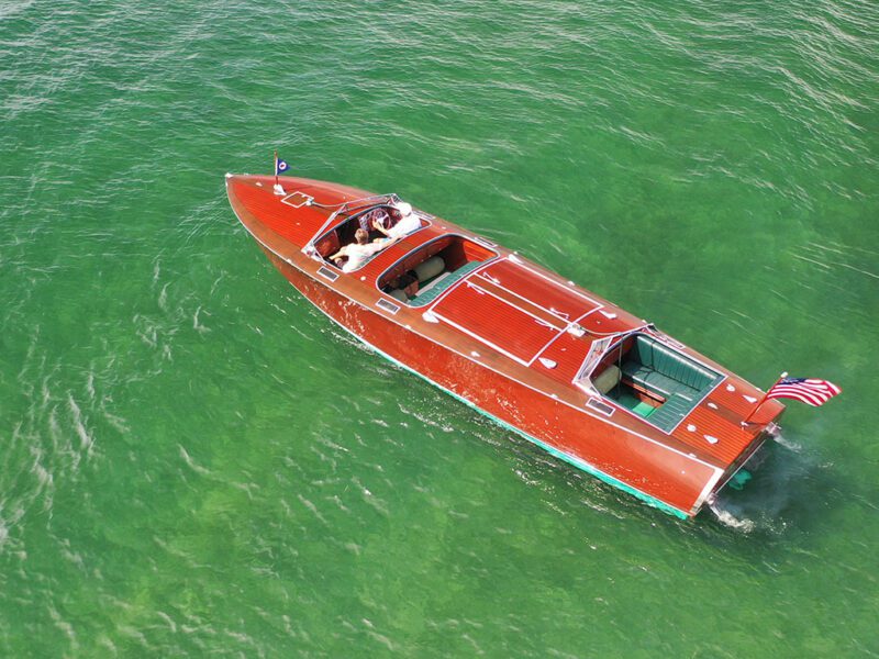 A vintage Mahogany Wooden Boat with an American flag glides across clear green water, carrying several passengers in classic Grand Prix Boat style.