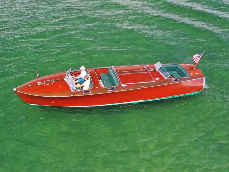A person drives a mahogany wooden boat with green upholstery on clear green water, an American flag displayed at the stern.