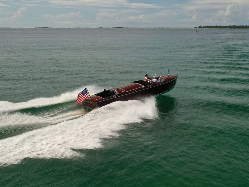 A mahogany wooden boat with an American flag moves quickly across calm green water, leaving a white wake behind. One person is seated at the helm, giving this vintage boat for sale a timeless elegance.