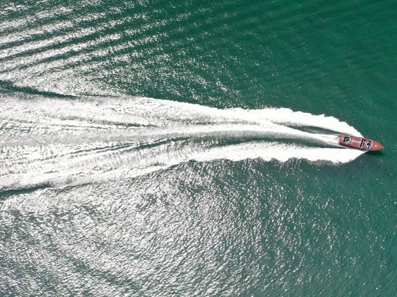 Aerial view of a sleek mahogany wooden boat speeding across green water, leaving a white wake behind it.
