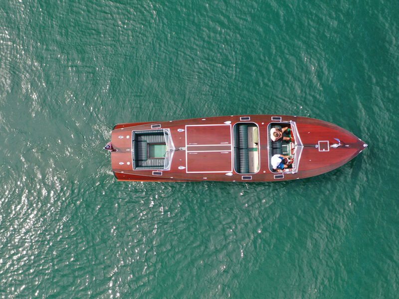 Aerial view of a mahogany wooden boat with two people on board, floating on calm green water.