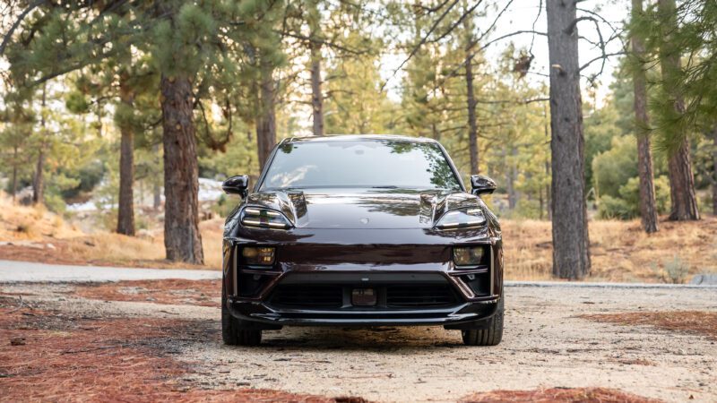 A dark-colored car is parked on a dirt path in a forested area with tall trees and scattered pine needles.