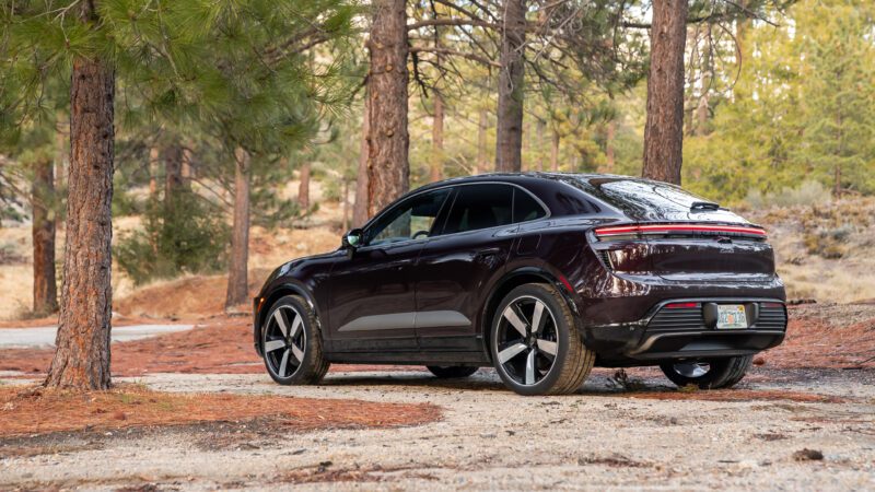 A dark-colored electric SUV is parked on a dirt path in a forested area with tall trees and scattered pine needles.