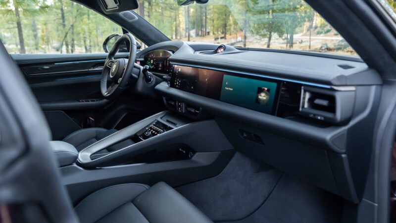 Modern car interior with black leather seats, digital dashboard displays, and touchscreen controls; forest scenery visible through the windows.