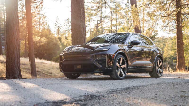 A black electric SUV is parked on a paved road in a forested area with sunlight filtering through the trees.