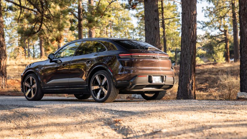 A dark-colored electric SUV is parked on a dirt road in a forested area with sunlight filtering through the trees.