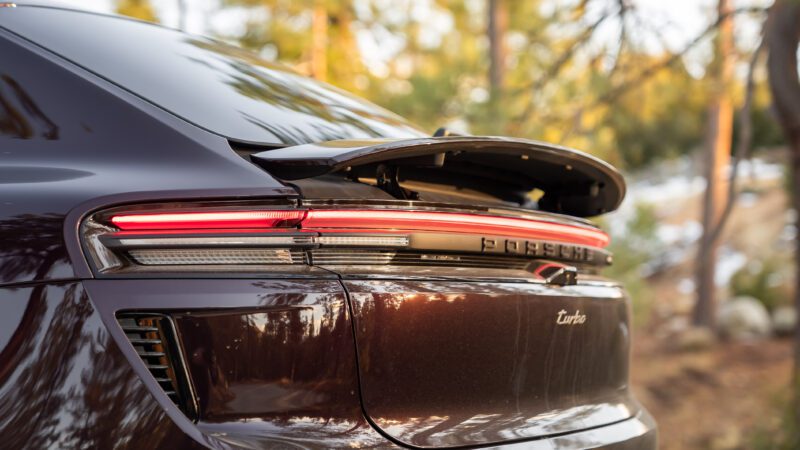 Close-up of the rear end of a dark-colored Porsche Taycan Turbo with its rear spoiler raised, parked outdoors among trees.