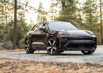 A dark-colored electric SUV is parked on a gravel road in a forested area with tall trees and scattered dry leaves.