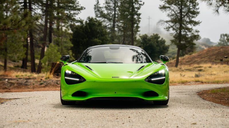 A bright green sports car with headlights on is parked on a rural road surrounded by trees and grass.