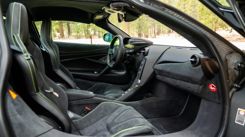 The interior of a modern sports car featuring black suede seats with green stitching, minimalist dashboard, and forest scenery visible through the windows.