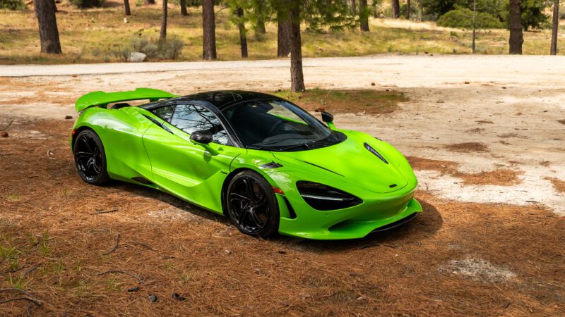 A bright green sports car parked on a pine needle-covered ground in a forested area with trees in the background.