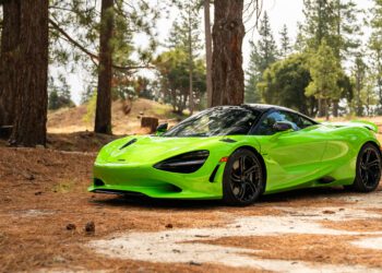 A bright green sports car is parked on a dirt path surrounded by tall pine trees in a forested area.