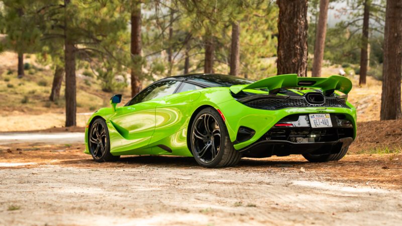 A bright green sports car is parked on a dirt path in a forested area with pine trees in the background.