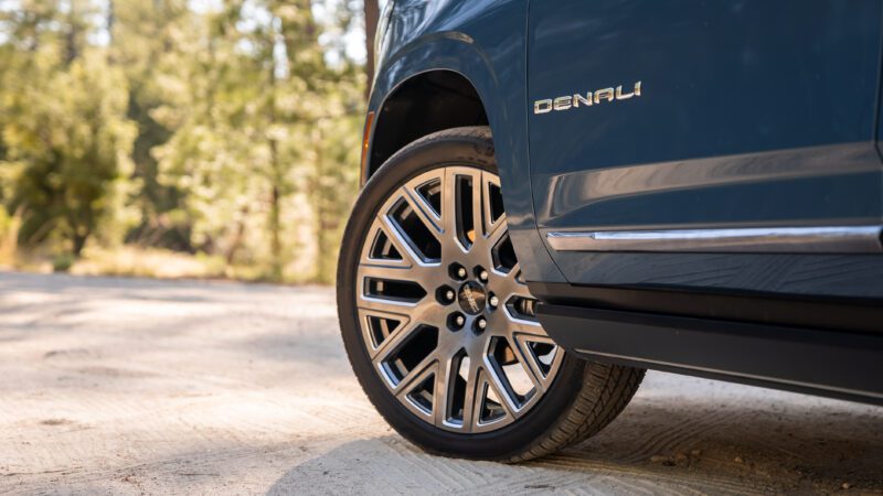 Close-up of a blue vehicle's front wheel and tire, featuring a multi-spoke alloy rim and "Denali" badge on the side, parked outdoors on a paved surface.