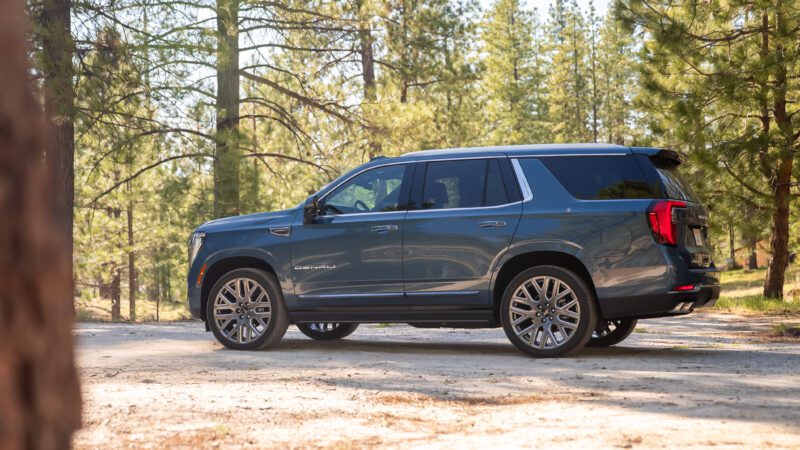 A blue GMC Yukon Denali SUV is parked on a dirt road in a forest with tall trees and sunlight filtering through the branches.