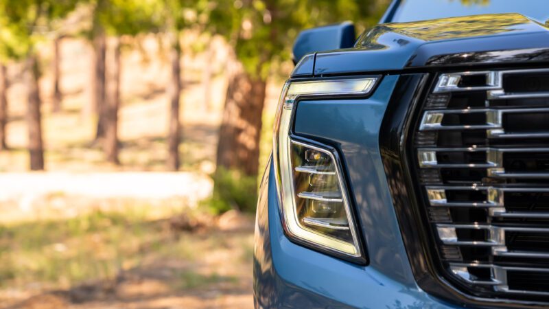 Close-up of the front left headlight and grille of a blue SUV, with trees and sunlight in the blurred background.