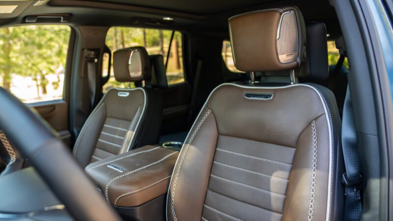 Front seats of a vehicle with brown leather upholstery, white stitching, and headrests, photographed from the driver’s side. Trees are visible outside the window.