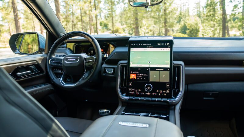 The interior of a modern SUV showing the steering wheel, large touchscreen infotainment system with navigation, and digital dashboard, with trees visible through the windshield.