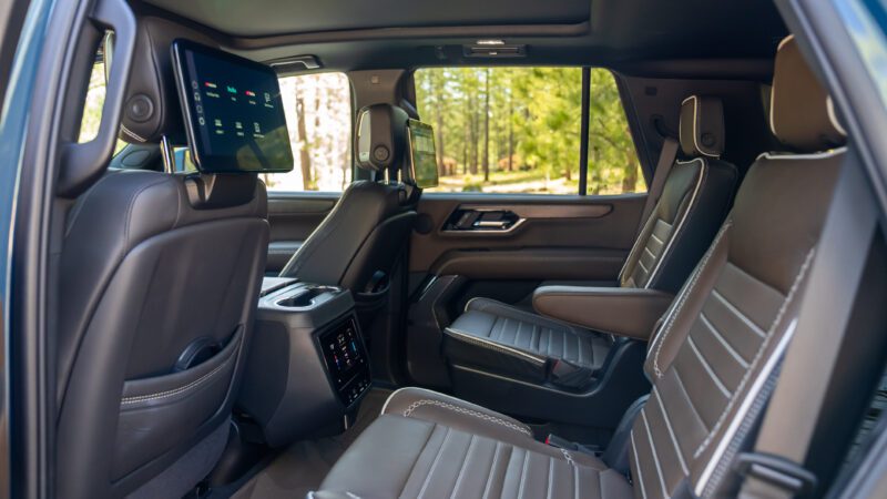 The interior of an SUV showing two rows of leather seats, rear-seat entertainment screens, and large windows with a view of trees outside.