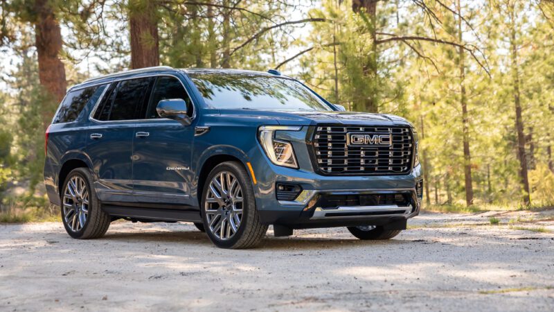 A blue GMC Yukon Denali SUV is parked on a dirt road in a forested area with tall trees in the background.