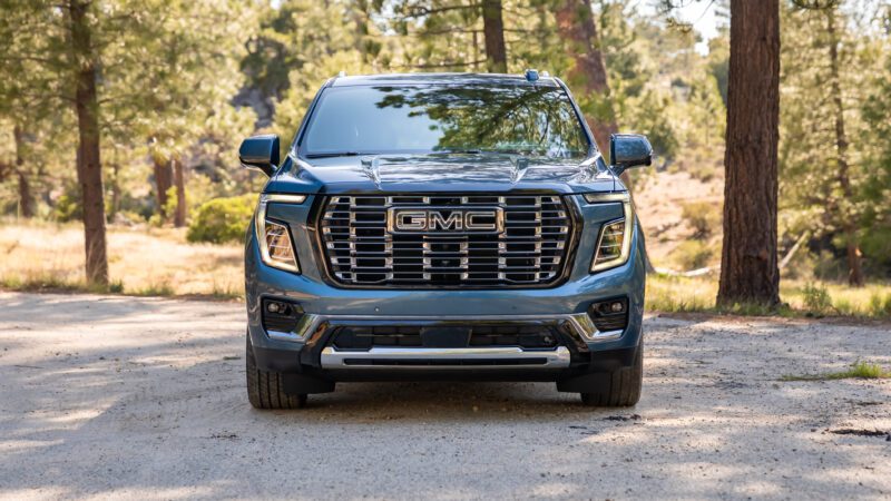 A blue GMC SUV is parked on a dirt road in a wooded area, shown from the front.