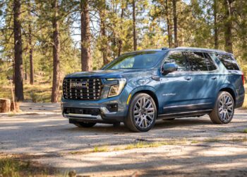 A blue GMC Yukon Denali SUV is parked on a dirt road in a wooded area with tall pine trees in the background.
