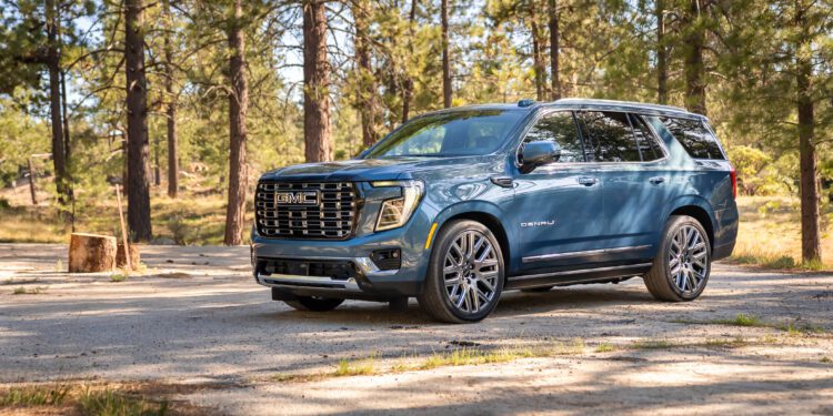A blue GMC Yukon Denali SUV is parked on a dirt road in a wooded area with tall pine trees in the background.