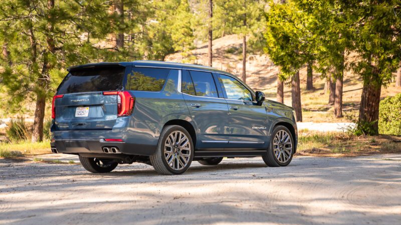 A blue GMC Yukon Denali SUV is parked on a dirt road in a wooded area with trees and sunlight in the background.