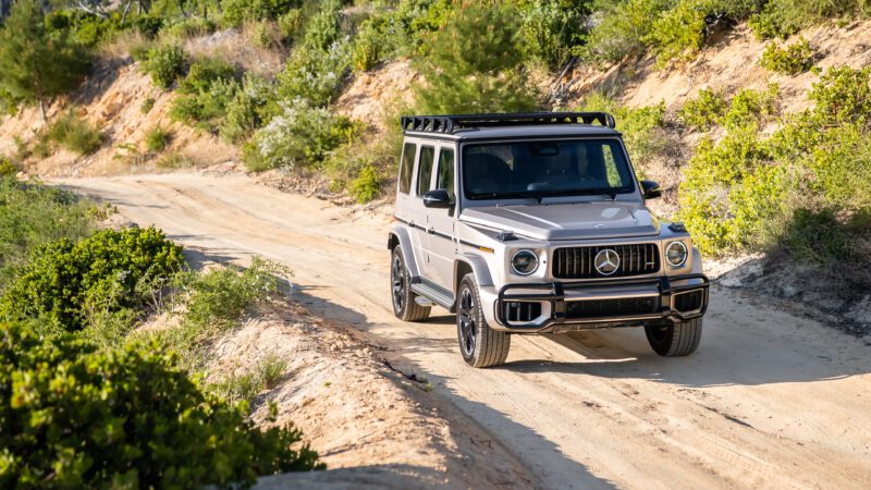 A silver Mercedes-Benz G-Class SUV drives on a dirt road surrounded by greenery and sandy terrain.
