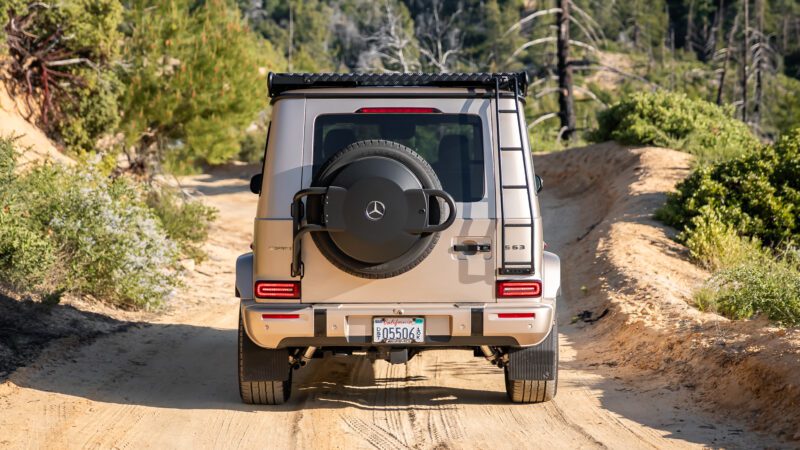 A beige Mercedes-Benz G-Class SUV drives on a dirt trail surrounded by trees and bushes, seen from the rear.