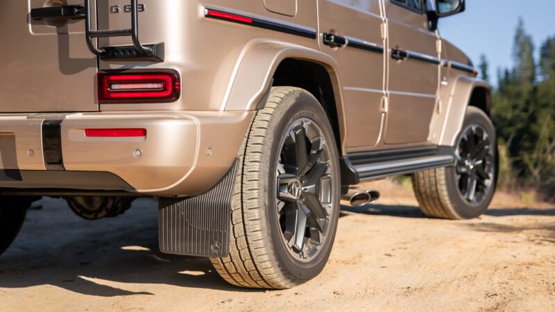 Close-up of the rear side of a tan Mercedes-Benz G-Class SUV parked on a dirt road, showing the wheel, taillights, and part of the bumper.