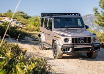 A silver Mercedes-Benz G-Class SUV with a roof rack is parked on a dirt road surrounded by greenery and mountains.