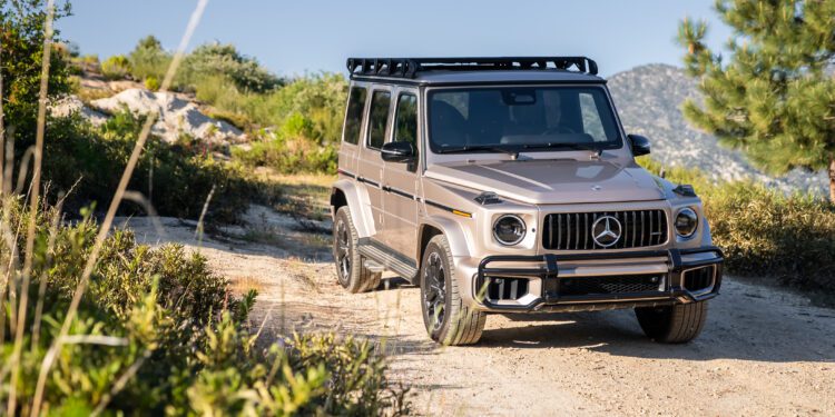 A silver Mercedes-Benz G-Class SUV with a roof rack is parked on a dirt road surrounded by greenery and mountains.