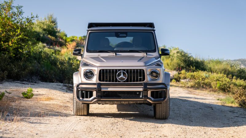 A silver Mercedes-Benz G-Class SUV is parked on a dirt road, surrounded by greenery under a clear blue sky.