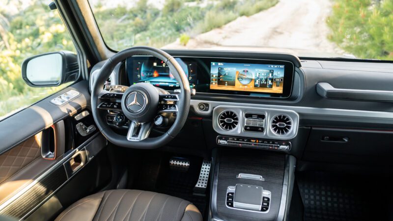 Interior of a Mercedes-Benz vehicle showing the steering wheel, digital dashboard, infotainment screens, air vents, and leather seats with a view of a dirt road outside.
