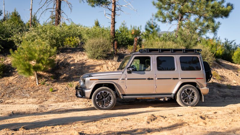 A beige SUV with black roof rack is parked on a dirt road surrounded by trees and vegetation under a clear sky.