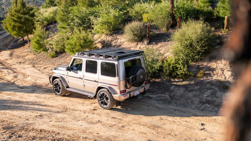 A silver Mercedes-Benz G-Class SUV drives on a dirt trail through a hilly, forested area on a sunny day.